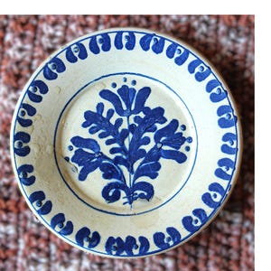 May include: A small, round, off-white ceramic plate with a blue floral design in the center. The rim features a repeating blue pattern. The plate is likely vintage or antique.