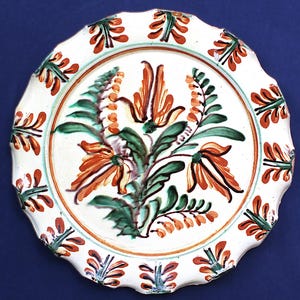 May include: White ceramic plate with a scalloped edge, featuring a floral design in orange, green, and brown. The plate has a central floral arrangement and repeating leaf patterns around the rim.