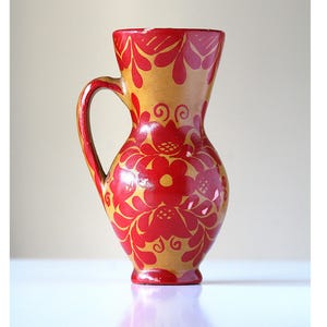 May include: A hand-painted, decorative pitcher with a handle. The pitcher is primarily tan with red floral designs. The red flowers and leaves are painted across the body and handle of the pitcher. The pitcher has a wide base and a narrow opening.