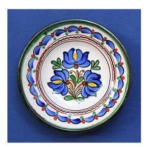 Puede incluir: Un plato pequeño y redondo de cerámica con base blanca y borde negro. El plato presenta un diseño floral central con tres flores azules con centros amarillos y hojas verdes. El borde está decorado con detalles azules y rojos.