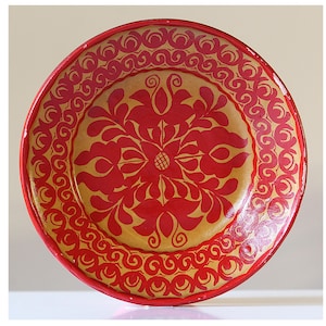 Könnte beinhalten: Ein runder, dekorativer Teller mit rotem Rand und goldenem Zentrum. Der Teller hat ein rotes Blumendesign in der Mitte und ein sich wiederholendes Wirbelmuster am Rand. Der Teller ist wahrscheinlich aus Keramik oder Porzellan.