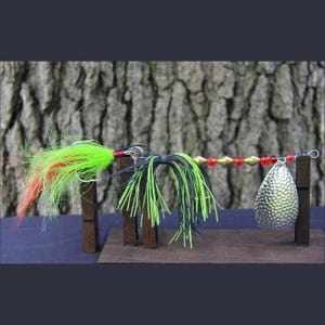 May include: A fishing lure with a silver, textured blade and a colorful feathered tail. The lure features red, green, and black accents, with a series of gold and red beads along the wire. It is displayed on a small wooden stand.