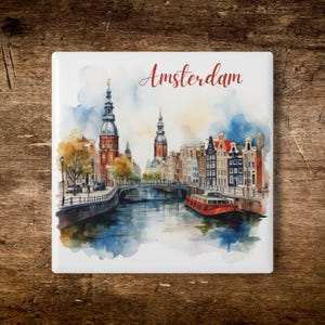 Op de afbeelding: Een vierkante keramische tegel met een aquarel schilderij van Amsterdam. De afbeelding toont een grachtenscène met gebouwen, bruggen en een rode boot. Het woord "Amsterdam" staat in rode letters bovenaan.