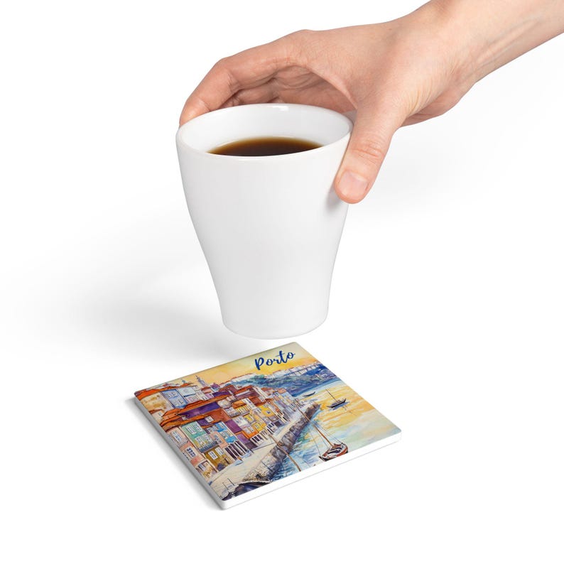 May include: A white ceramic coffee mug filled with dark liquid, held above a square coaster. The coaster features a watercolor painting of a coastal city with the word "Porto" in blue script.