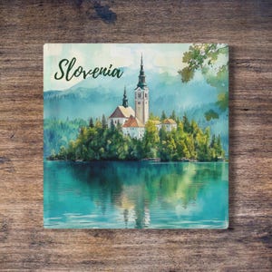 Può includere: Un dipinto ad acquerello del lago di Bled in Slovenia. L'immagine presenta una chiesa su un'isola circondata da alberi verdi, con la parola "Slovenia" in verde in alto. L'acqua del lago è di un vibrante colore turchese.