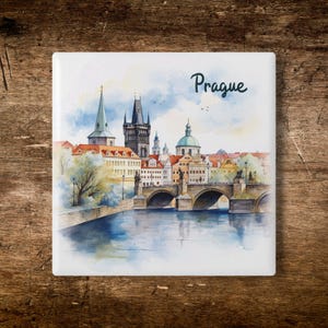 May include: Square ceramic tile featuring a watercolor painting of Prague, Czech Republic. The artwork depicts the Charles Bridge, buildings, and a river under a blue sky. The word "Prague" is written in cursive.