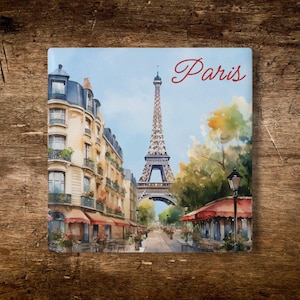 May include: A square decorative tile featuring a watercolor-style illustration of the Eiffel Tower in Paris. The scene includes classic Parisian buildings with balconies, red awnings, street lamps, and trees. The word Paris is written in red script.