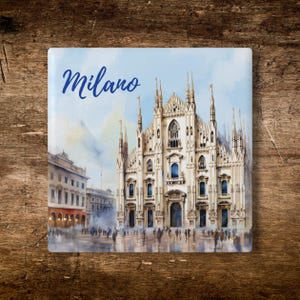 May include: A square ceramic tile featuring a watercolor illustration of the Duomo di Milano cathedral in Italy. The word Milano is written in blue script in the upper left corner. The scene depicts the gothic architecture and a public square.