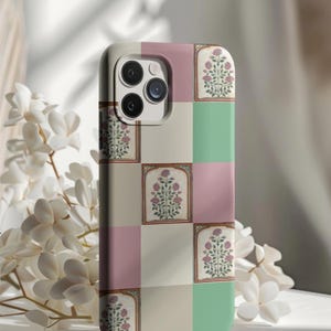 Op de afbeelding: Een telefoonhoesje met een geblokt patroon in roze, groen en crème. Elke vierkant heeft een omlijnd bloemenontwerp met roze bloemen. De hoes heeft een camera-uitsparing en staat tegen een zachte, vage achtergrond met witte bloemen.