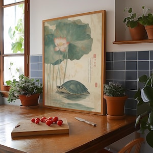 May include: Framed artwork featuring a turtle in water, with a lotus flower and leaves. The artwork has a light brown wooden frame. The background is a light beige color. The artwork is displayed on a wooden surface.