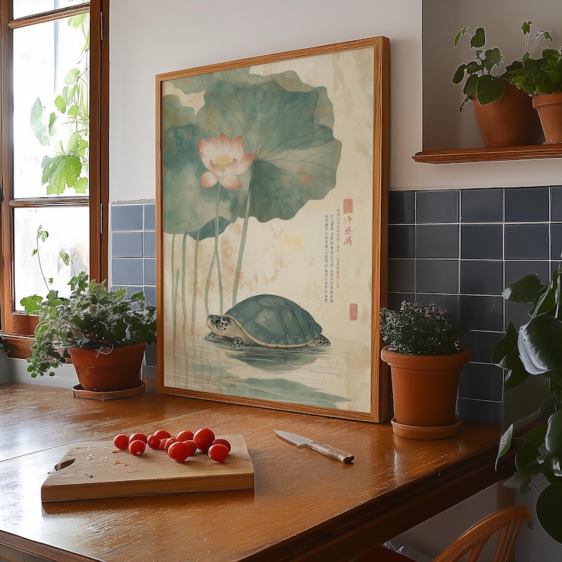 May include: Framed artwork featuring a turtle in water, with a lotus flower and leaves. The artwork has a light brown wooden frame. The background is a light beige color. The artwork is displayed on a wooden surface.