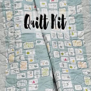 Op de afbeelding: Een lichtblauwe quiltkit met een herhalend patroon van geïllustreerde vierkanten, letters en woorden. De tekst "Quilt Kit" wordt weergegeven in grote zwarte letters. De quilt heeft een lichtblauwe rand met een stippenpatroon.