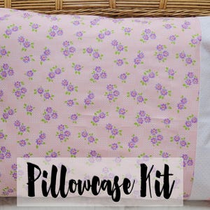 May include: Pink pillowcase kit with a floral pattern of small purple flowers and green leaves. The pillowcase has a white and pink polka dot border and the text "Pillowcase Kit".