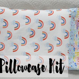 Pode incluir: Um kit de fronha com uma fronha branca com um padrão de arco-íris e uma fronha azul com um padrão de texto colorido. O texto na fronha azul diz "Shine Love Stay Wonder Sun Shine". As fronhas são etiquetadas como "Pillowcase Kit".