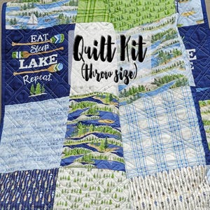 Op de afbeelding: Een patchwork quilt kit in werpgrootte, met verschillende stofpatronen met een meer-thema. De quilt bevat blauwe, groene en witte stoffen met afbeeldingen van boten, bomen en de tekst "Eat, Sleep, Lake, Repeat".