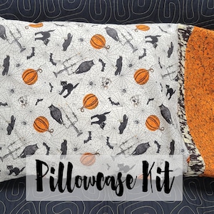 May include: A pillowcase kit featuring a white fabric with a black and orange Halloween print. The print includes bats, owls, pumpkins, spiders, and cats. The pillowcase is trimmed with a black and orange fabric. The text "Pillowcase Kit" is printed on the front of the pillowcase.