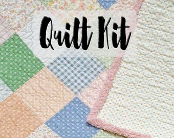 May include: A patchwork quilt kit with a variety of pastel fabrics, including floral, polka dot, and gingham patterns. The quilt is bordered with a pink, ruffled edge. The words "Quilt Kit" are displayed in a bold, black font.
