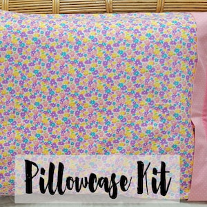 Op de afbeelding: Een kussensloopset met een bloemenpatroon in roze, geel en blauw. De set bevat een kussensloop met een bloemenontwerp en een effen roze kussensloop. De tekst "Pillowcase Kit" staat op de voorkant.