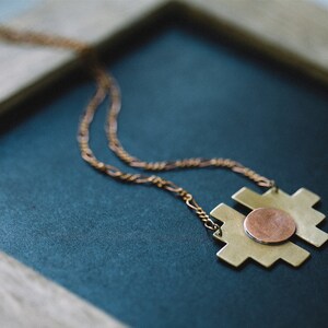 Op de afbeelding: Een gouden ketting met een geometrische hanger met een koperen cirkel in het midden. De ketting wordt getoond op een donkerblauw oppervlak.