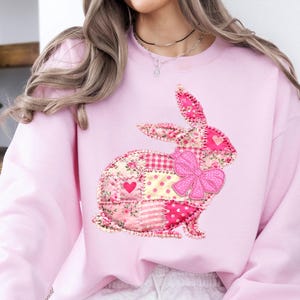 May include: A pale pink sweatshirt with a patchwork bunny design and a large pink bow. The bunny is made from various pink patterned fabrics, including hearts and polka dots. The sweatshirt is a soft, comfortable style.