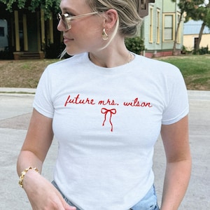 May include: White t-shirt with the text "future mrs. wilson" in red script and a red bow graphic. The shirt is short-sleeved and worn with blue jeans. The person is wearing gold jewelry and sunglasses.