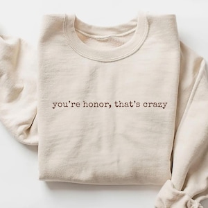 May include: Sand-colored sweatshirt with the phrase "you're honor, that's crazy" printed in brown. The sweatshirt has a crew neck and long sleeves, with the sleeves cuffed. The word "Sand" is written in cursive at the bottom left.