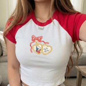 May include: White and red short-sleeved crop top with a graphic design. The top features a white body with red sleeves and collar. The graphic includes two heart-shaped lockets with a cartoon character and the text "Some Imagination Huh?"