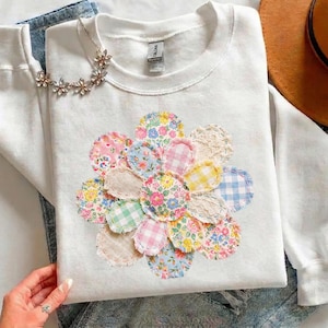 May include: White sweatshirt featuring a large, colourful floral appliqué made from various patterned fabrics. A delicate floral necklace rests on the sweatshirt. The image also includes a brown hat and denim jeans.
