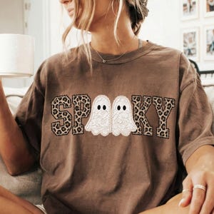 May include: A brown short-sleeve t-shirt featuring the text SPOOKY in a leopard print pattern, with two white lace-textured ghost illustrations replacing the letter O. The shirt has a crew neckline and a relaxed fit, showcasing a graphic design.