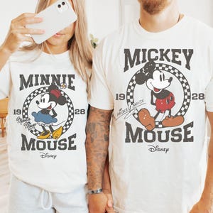 Puede incluir: Camisetas blanquecinas con Mickey y Minnie Mouse. Cada camiseta tiene un gr&aacute;fico circular con la imagen del personaje, el texto "MICKEY MOUSE" o "MINNIE MOUSE" y el logotipo de Disney. Las camisetas se combinan con pantalones cortos grises.