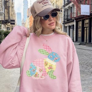 May include: Pale pink sweatshirt with a patchwork bunny design in floral and gingham patterns. The bunny is in shades of pink, blue, green, and yellow. The sweatshirt is worn with a baseball cap that says "The Western Casino Las Vegas Nevada".