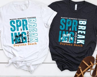 Op de afbeelding: Twee T-shirts, één witte en één houtskoolkleurige, met een strandthema. De shirts lezen "Spring Break 2026 Daytona Beach" in teal en oranje. Bruine sandalen zijn geplaatst in de buurt van het houtskoolkleurige shirt.