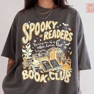 May include: A charcoal grey short-sleeve t-shirt featuring a graphic print with the text "SPOOKY READERS BOOK CLUB" and "Running on coffee, horror & chapters". The design includes illustrations of books, a skull, candles, a pumpkin, and plants.