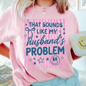 May include: A light pink t-shirt with the text "THAT SOUNDS LIKE MY husband's PROBLEM" in blue and pink. The shirt also features stars and a smiley face. A white mug with a yellow smiley face is held in the image.
