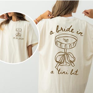 May include: Cream-colored t-shirt with a brown graphic of a martini glass and the phrase "a bride in a tini bit." The front of the shirt features a smaller martini glass graphic and the date "11.10.2025."