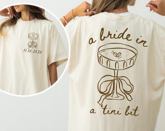May include: Cream-colored t-shirt with a brown graphic of a martini glass and the phrase "a bride in a tini bit." The front of the shirt features a smaller martini glass graphic and the date "11.10.2025."