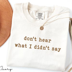 May include: Ivory-colored t-shirt with the text "don't hear what I didn't say" in brown lettering. A gold necklace with a pendant and a brown hat are also visible. The shirt has a Comfort Colors tag.