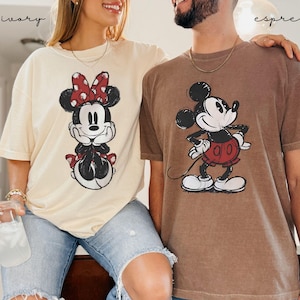 Puede incluir: Dos camisetas de manga corta, una marfil y otra espresso, con gr&aacute;ficos de Mickey y Minnie Mouse. La camiseta de Minnie Mouse es marfil, y la camiseta de Mickey Mouse es espresso. Ambas camisetas tienen un aspecto vintage y desgastado, con los personajes en blanco y negro.