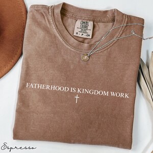 May include: Espresso-colored t-shirt with the text "FATHERHOOD IS KINGDOM WORK" and a cross. The shirt has a crew neck and short sleeves. A silver necklace with a pendant is draped across the shirt.