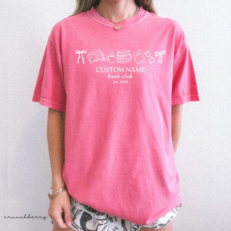 May include: A bright pink short-sleeved t-shirt featuring white line art graphics of bows, books, a coffee mug, and glasses. The text reads CUSTOM NAME book club est. 2026. The shirt has a crew neckline and a relaxed fit, showcasing a casual style.