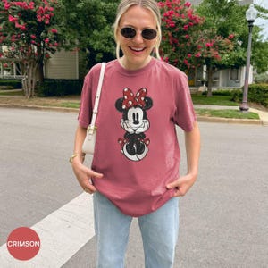 Puede incluir: Camiseta color carmes&iacute; con un gr&aacute;fico de Minnie Mouse, lazo rojo y lunares. La camiseta se combina con vaqueros azul claro. La palabra "CRIMSON" est&aacute; en un c&iacute;rculo rojo en la esquina inferior izquierda.