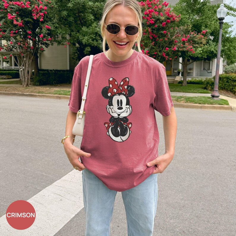 Puede incluir: Camiseta color carmes&iacute; con un gr&aacute;fico de Minnie Mouse, lazo rojo y lunares. La camiseta se combina con vaqueros azul claro. La palabra "CRIMSON" est&aacute; en un c&iacute;rculo rojo en la esquina inferior izquierda.