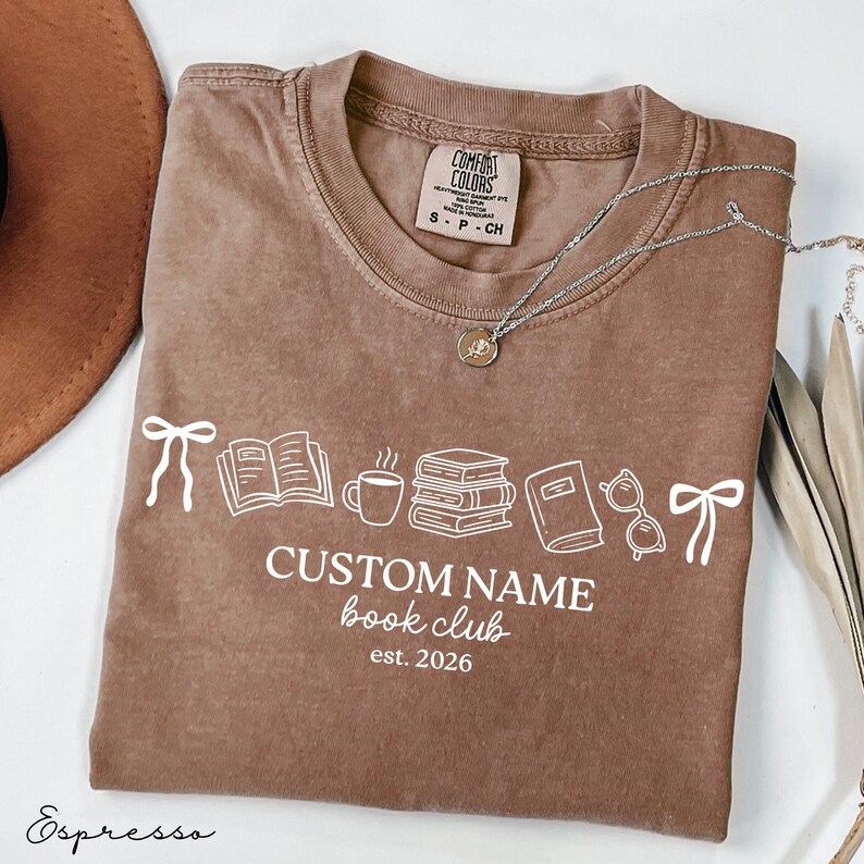 May include: A brown short-sleeve t-shirt featuring a white graphic print of two bows, an open book, a coffee mug, a stack of books, a closed book, and glasses. The text reads CUSTOM NAME book club est. 2026. The shirt has a Comfort Colors brand label.