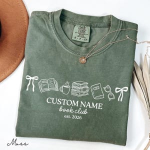 May include: A moss green t-shirt featuring a white graphic print of two bows, an open book, a coffee mug, a stack of books, a closed book, and eyeglasses. Below the graphics, the text reads CUSTOM NAME book club est. 2026 in white lettering.