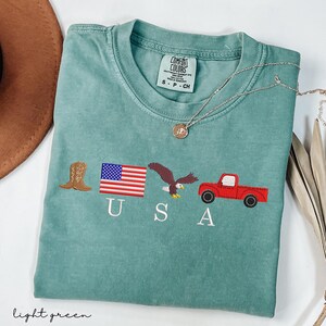 May include: Light green t-shirt with embroidered patriotic symbols: a cowboy boot, American flag, eagle, and red truck. The word "USA" is embroidered below the symbols. The shirt has a Comfort Colors tag.