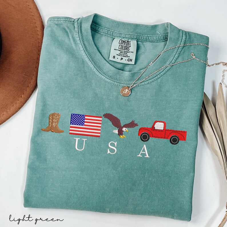 May include: Light green t-shirt with embroidered patriotic symbols: a cowboy boot, American flag, eagle, and red truck. The word "USA" is embroidered below the symbols. The shirt has a Comfort Colors tag.