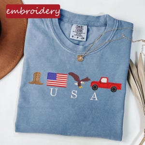 May include: A light blue Comfort Colors t-shirt with embroidered patriotic designs. The design includes a brown cowboy boot, a US flag, an eagle, a red truck, and the letters "USA". A gold necklace is draped across the shirt. The word "embroidery" is in a red box.