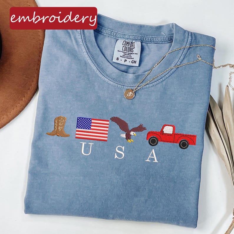 May include: A light blue Comfort Colors t-shirt with embroidered patriotic designs. The design includes a brown cowboy boot, a US flag, an eagle, a red truck, and the letters "USA". A gold necklace is draped across the shirt. The word "embroidery" is in a red box.