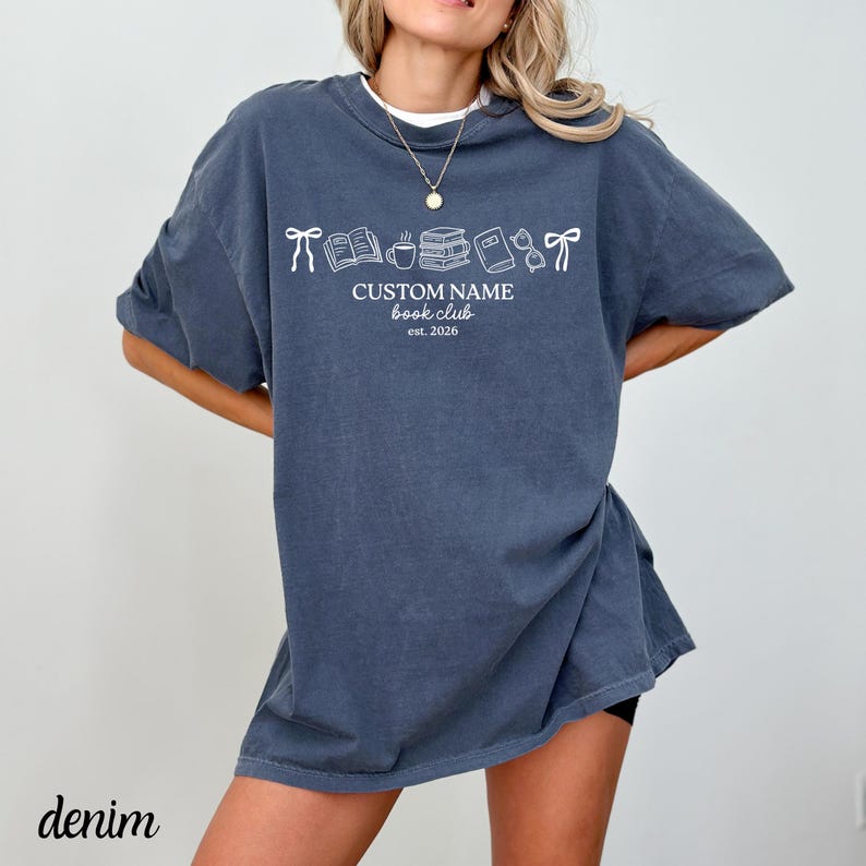 May include: A slate blue short-sleeve t-shirt featuring a white graphic print of bows, books, a coffee mug, and glasses. The text reads CUSTOM NAME book club est. 2026. The shirt has a crew neckline and a relaxed, loose-fitting silhouette.