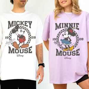 Puede incluir: Dos camisetas holgadas, una blanca y otra lavanda, con dise&ntilde;os de Mickey y Minnie Mouse. La camiseta blanca muestra a Mickey Mouse con el texto "Mickey Mouse" y "1928". La camiseta lavanda muestra a Minnie Mouse con el texto "Minnie Mouse" y "1928".